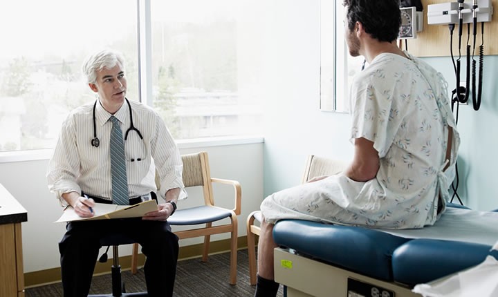 Medico sentando e uma escutando os relatos de sua paciente que está sentada em uma cama.